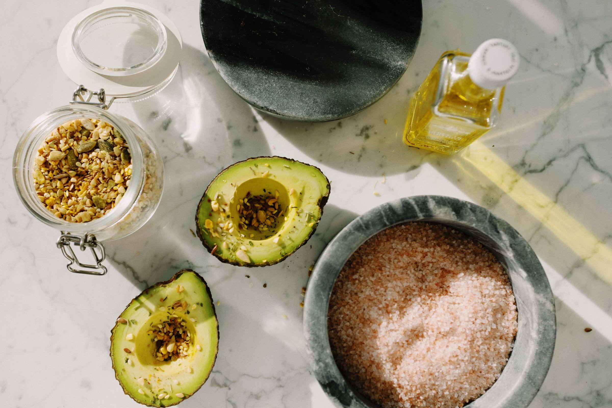 Halbierte Avocados mit Saatenmischung, ein Glas Kerne, rosa Salz und Olivenöl auf Marmor