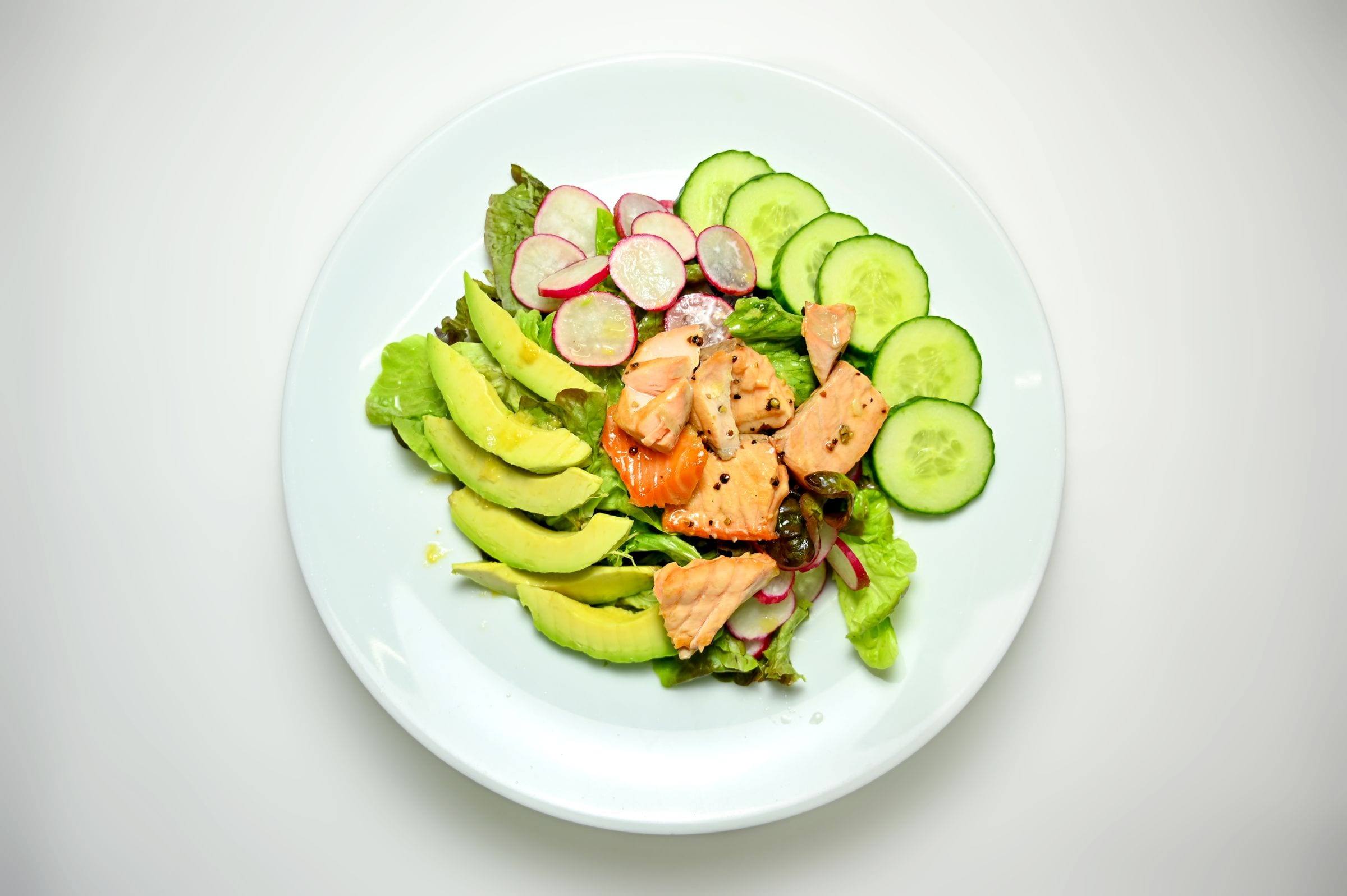 Salat mit Lachs, Avocadoscheiben, Gurke und Radieschen auf einem weißen Teller